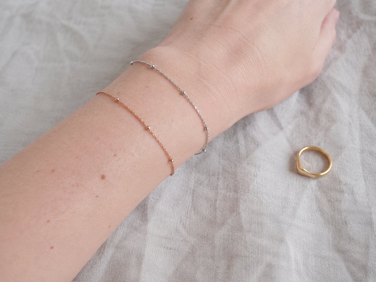 Pols met twee fijne armbanden in zilver en roségoud, met een klein gouden ringetje op een beige stof