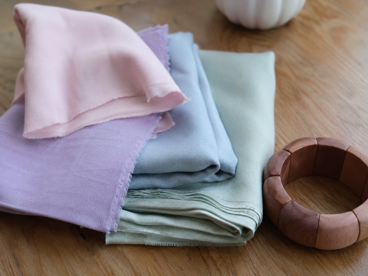 Gevouwen stofstalen in pastelkleuren roze, lila, lichtblauw en saliegroen op een houten ondergrond met een houten armband